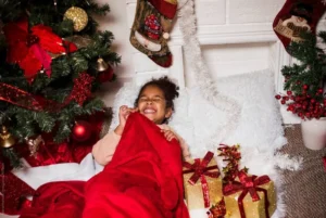 girl smiling under a red blanket with Christmas presents by a festive tree.