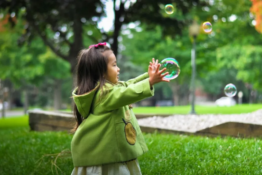 Little girl in a green coat playing with bubbles outdoors.