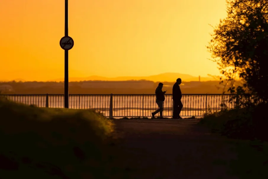 Silhouette of two people walking against a sunset sky near a railing
