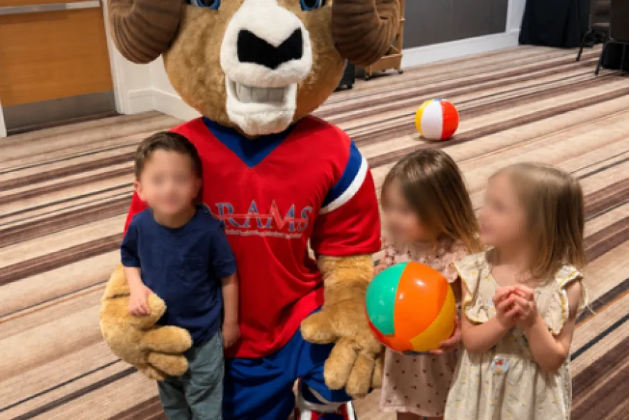 Children posing with a ram mascot holding colorful balls in an indoor play area.