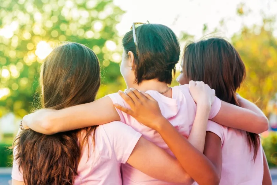 Three people embracing while standing outdoors in matching pink shirts on a sunny day.