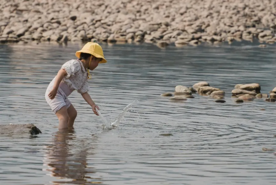Child wearing a yellow hat playing in a shallow river with rocks in the background