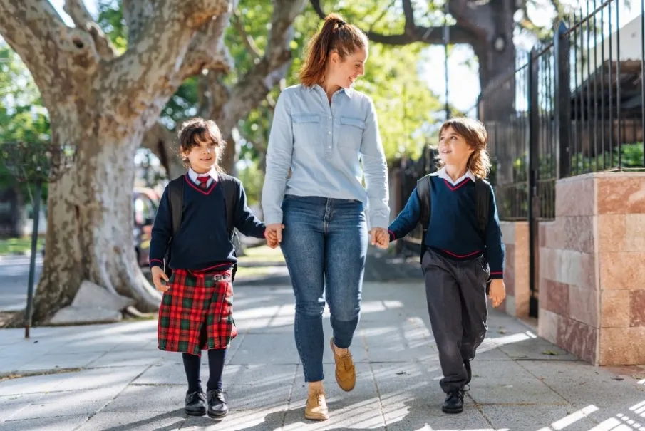 Mother walking with two children in school uniforms on a tree-lined sidewalk