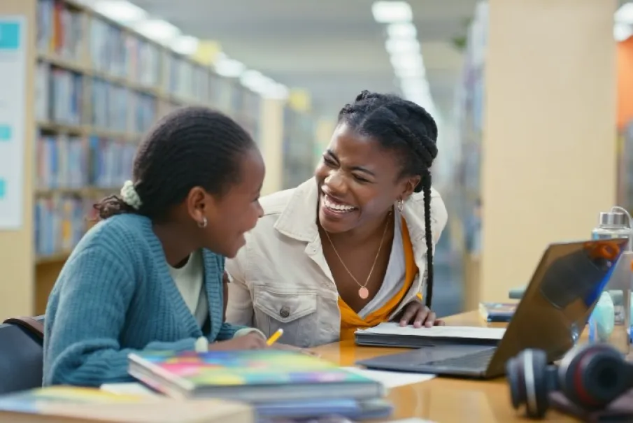 Teacher and student smiling in a library with books and a laptop