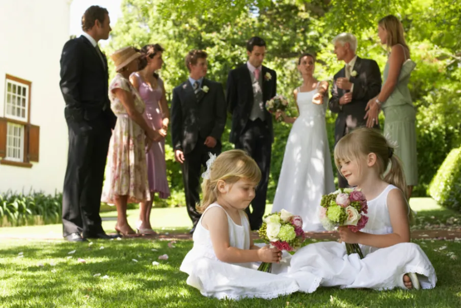Two young girls in white dresses holding bouquets at a wedding with guests in the background