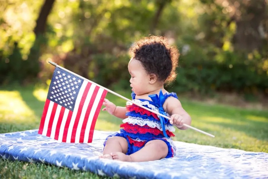 Baby sitting on a picnic blanket holding an American flag in a park.