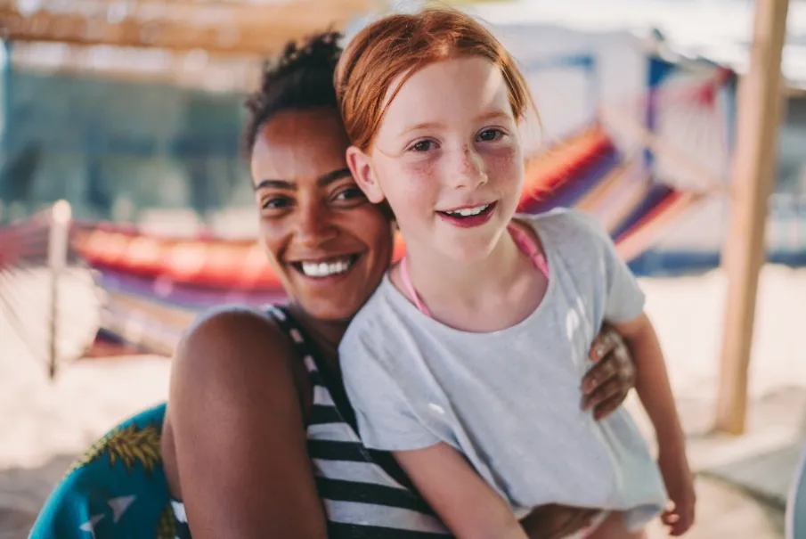 Smiling woman hugging a child at the beach with a hammock in the background
