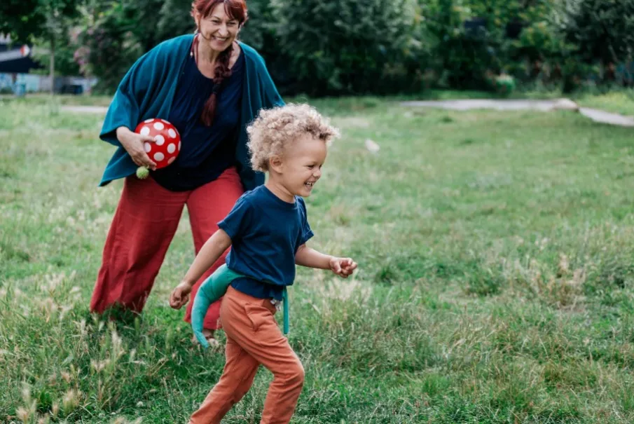 Child running in the park with woman holding red spotted ball.
