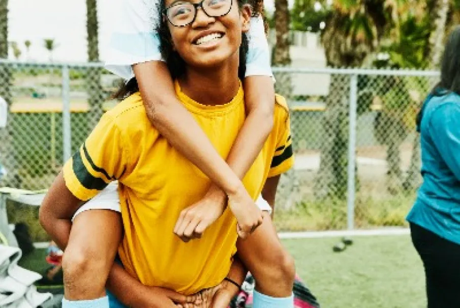 Two friends laughing while giving a piggyback ride at an outdoor sports field.