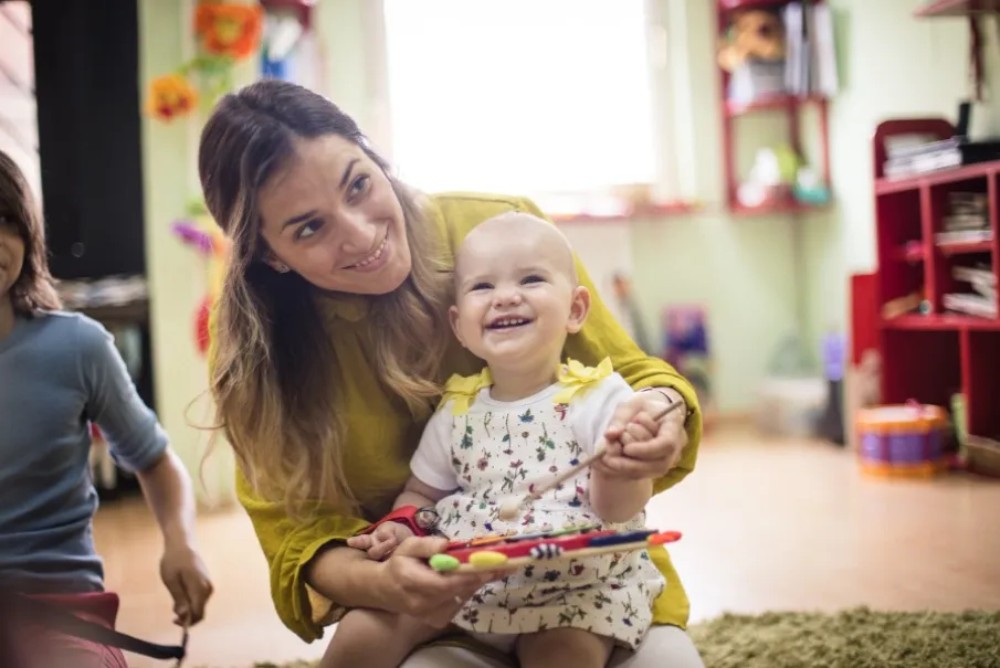 Woman smiling at baby holding a tablet in a colorful daycare setting