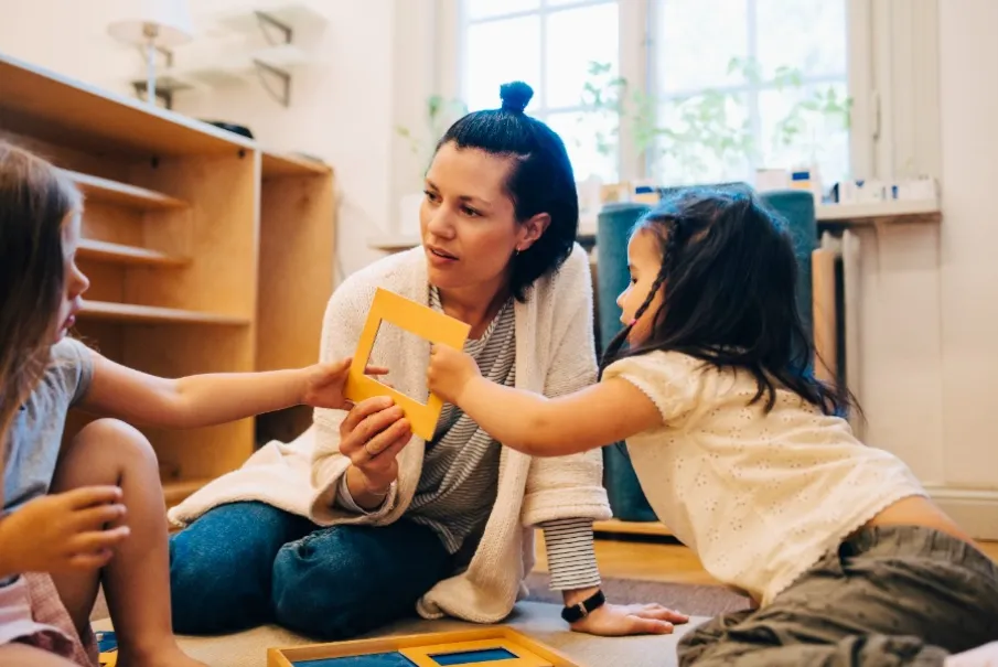 Teacher interacting with two young children using educational wooden shapes in a classroom setting.