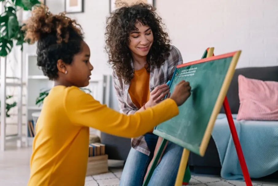 Child learning to write with tutor assistance at a chalkboard.