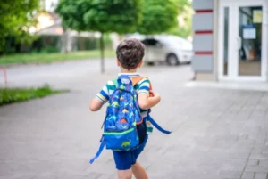 Young boy wearing a dinosaur backpack walking on a sidewalk towards a building.