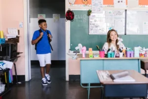 Student entering classroom with teacher smiling at desk