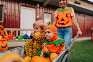 Children in tiger and pumpkin costumes sitting on a wheelbarrow with pumpkins during Halloween.