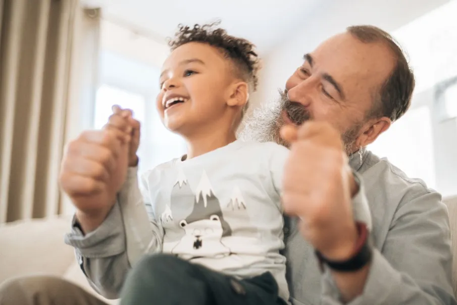 Older man with a beard playing joyfully with a young boy in a bright living room.