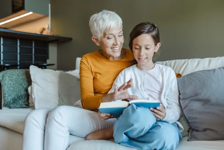 Grandmother reading book with granddaughter on cozy living room couch.