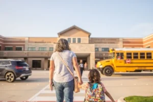 Parent and child with a colorful backpack approaching a school building next to a yellow school bus.