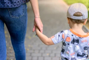 Child in graphic shirt and cap holding an adult's hand while strolling on a paved walkway.