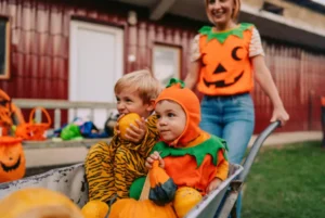 Children dressed in pumpkin costumes sitting in a wheelbarrow with pumpkins.