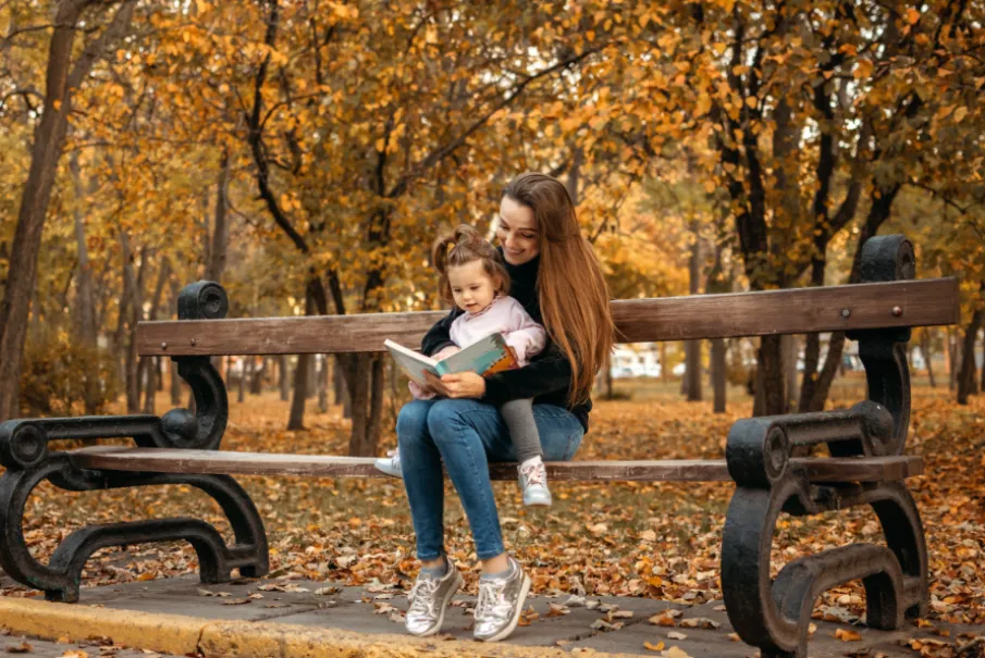 Mother and daughter reading on a park bench surrounded by autumn leaves