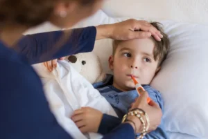Child in bed with a thermometer and hand on forehead checking temperature