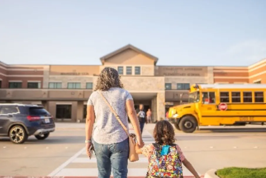 Mother and child walking toward school building with a school bus parked nearby