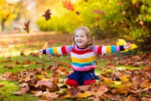 Happy child playing with falling leaves in the park during colorful autumn day