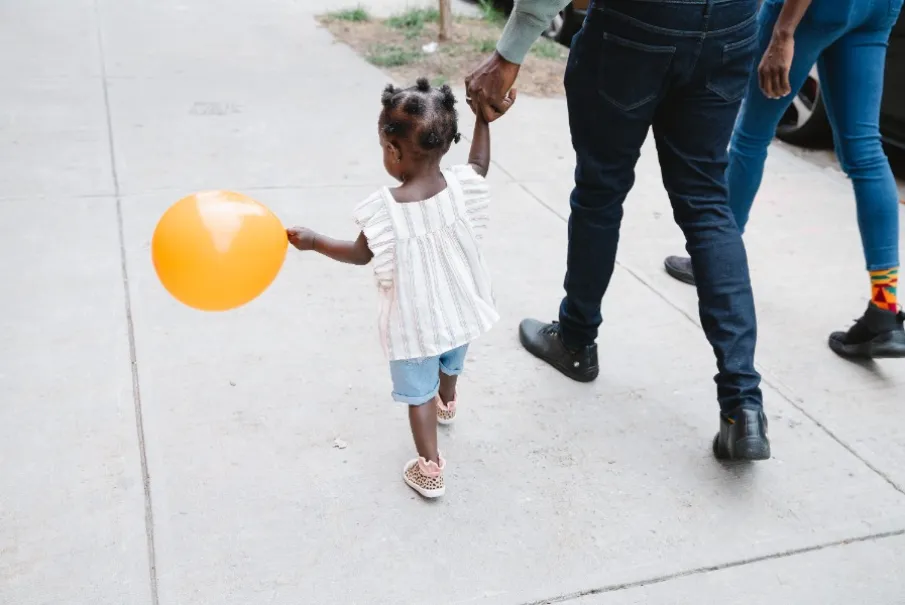 Toddler holding orange balloon while walking hand in hand with an adult on a sidewalk