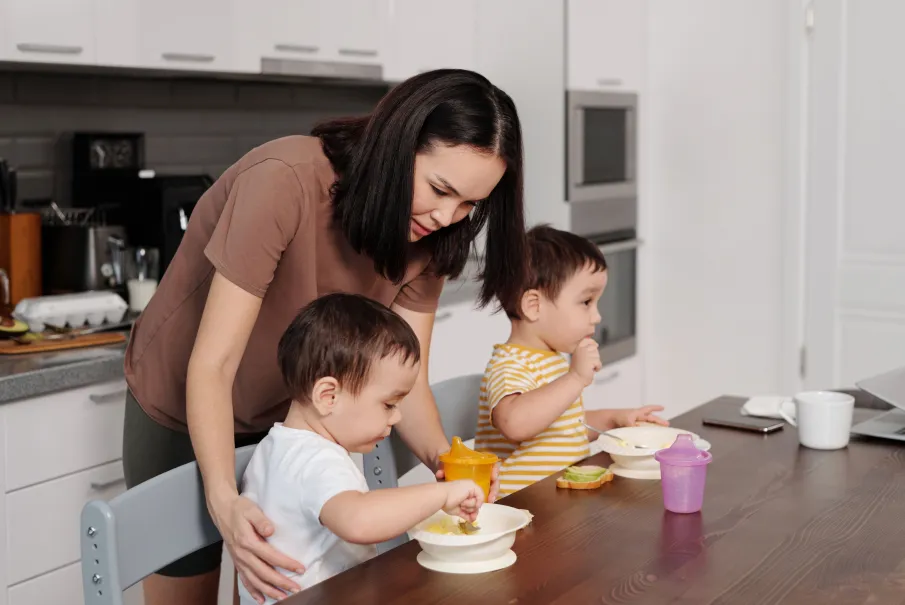 Mother helping two young children eat at a kitchen table.
