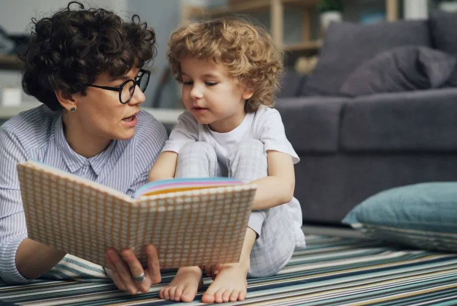 Woman reading a book to a young child on a striped rug in a comfortable home setting.